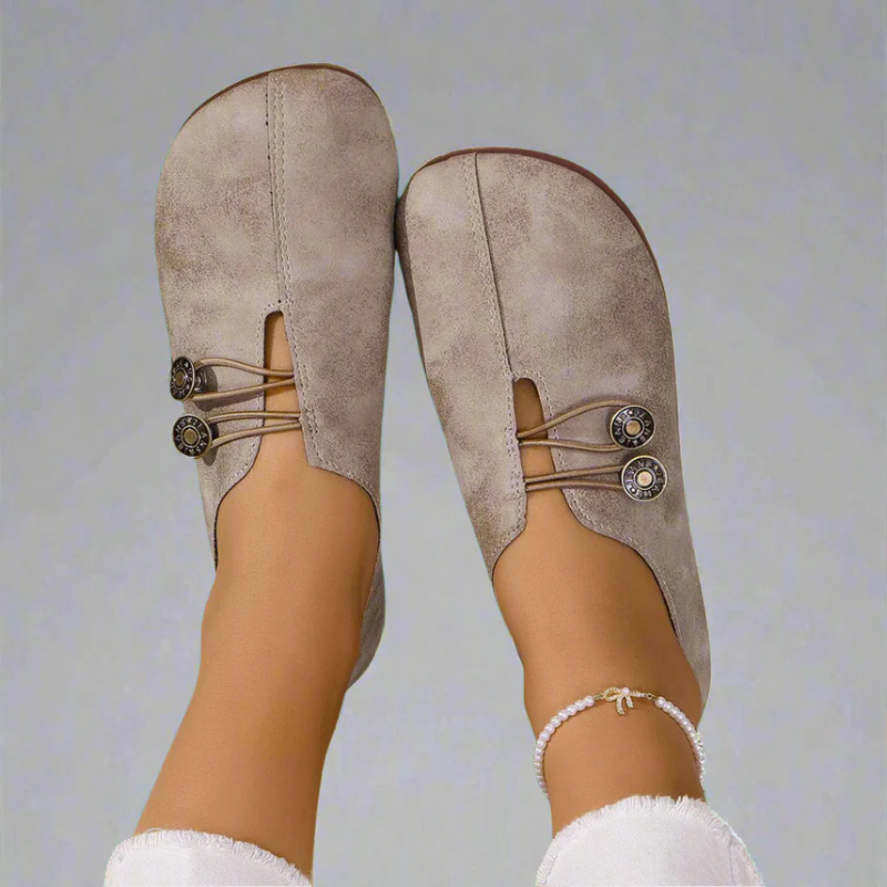 Beige Damen Mokassins mit dekorativen Knöpfen und elastischen Bändern, getragen mit weißer Hose und Perlenfußkettchen. Bequeme Freizeitschuhe.