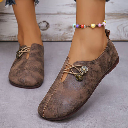 Braune Damen-Loafers mit Knopfdetails und bunter Perlenfußkette, auf grauem Boden. Modische, bequeme Schuhe für den Alltag.