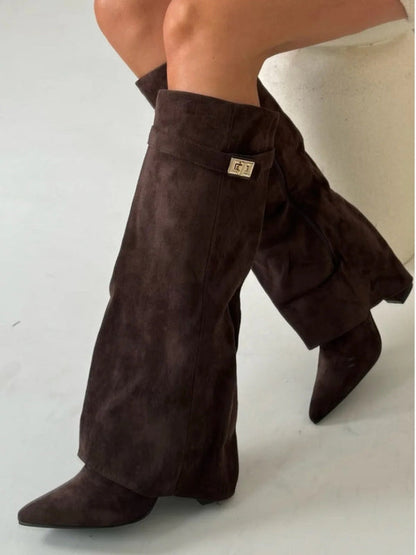 Dunkelbraune Wildlederstiefel mit spitzer Zehenpartie und goldener Schnalle, getragen von einer Person, die auf einem weißen Hocker sitzt.