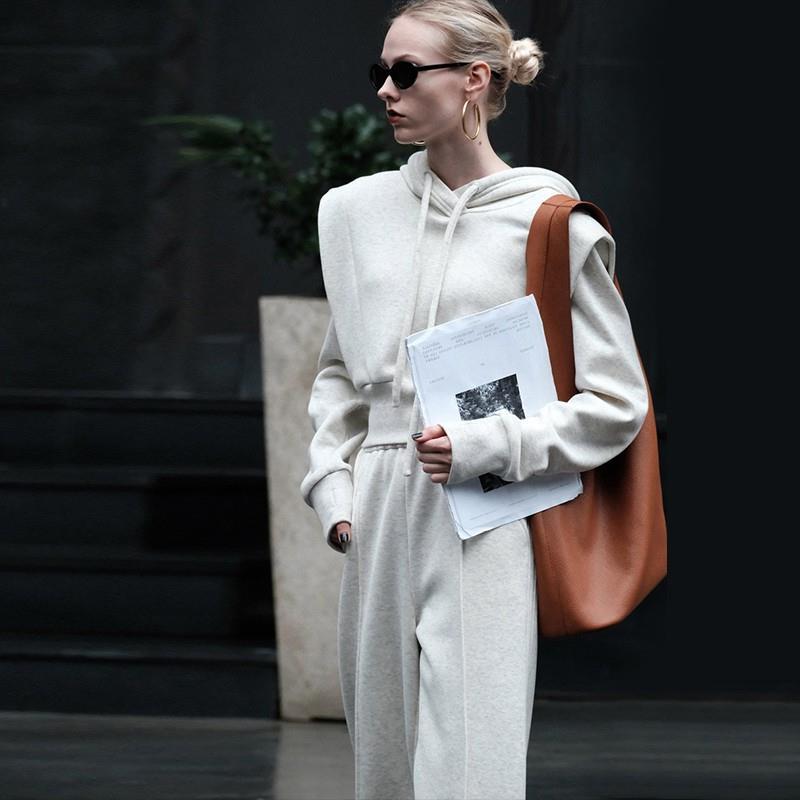 Frau in grauem Hoodie und Jogginghose, trägt braune Tasche und Dokumente, modisch, urbaner Stil, Sonnenbrille, moderne Streetwear.