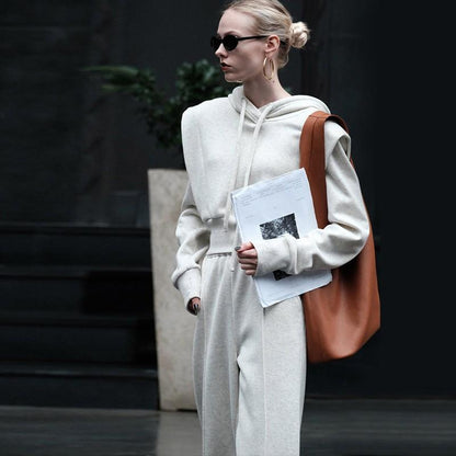 Frau in grauem Hoodie und Jogginghose, trägt braune Tasche und Dokumente, modisch, urbaner Stil, Sonnenbrille, moderne Streetwear.