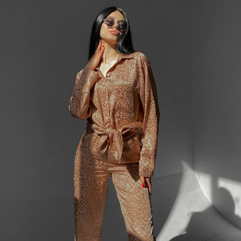 Frau in eleganter, beiger Pyjama-Set mit Leopardenmuster, Sonnenbrille und langen schwarzen Haaren posiert vor grauem Hintergrund. Fashion, Nachtwäsche.