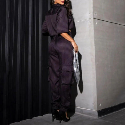 Frau in schwarzem, gestreiftem Jumpsuit mit silberner Clutch und High Heels, steht vor einer modernen, dunklen Wand. Elegante Abendmode.