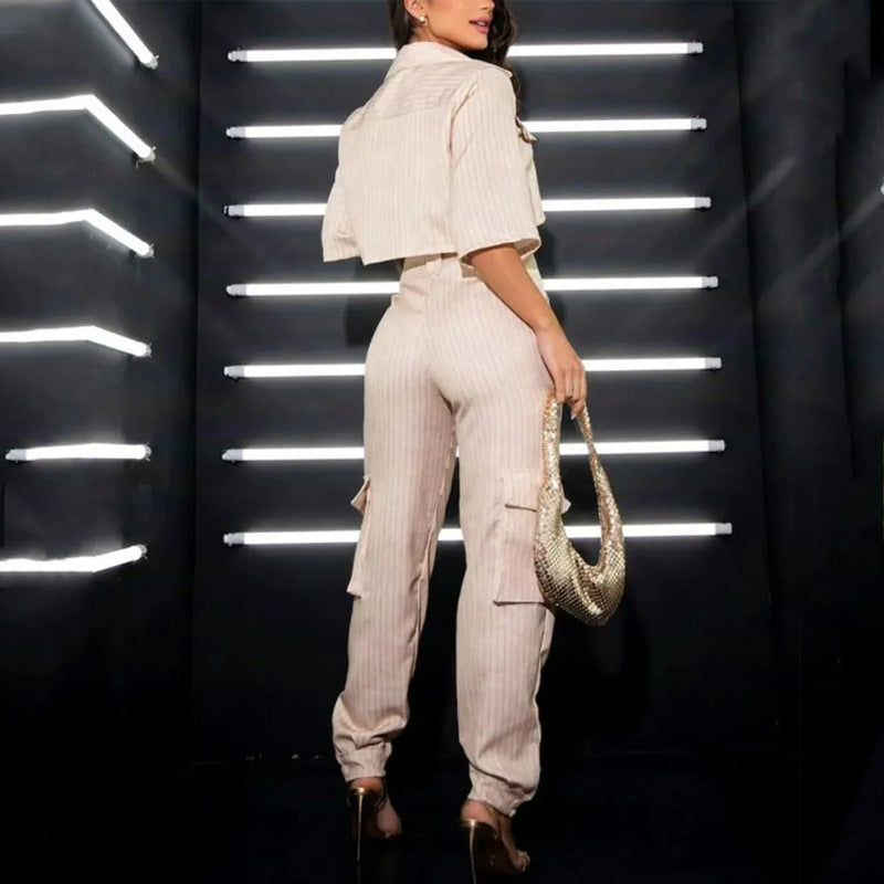 Frau in beigem, gestreiftem Jumpsuit mit goldener Tasche, posiert vor moderner LED-Leuchtenwand. Elegante Mode, stilvolle Beleuchtung.