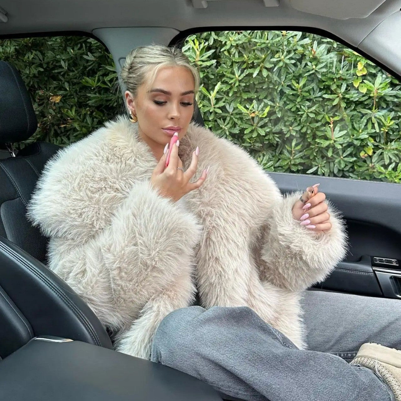 Frau im Auto mit beigem Kunstpelzmantel, hält Lippenstift. Elegante Mode, luxuriöser Stil, Herbst/Winter-Trend, moderne Frau, Autofahrt.