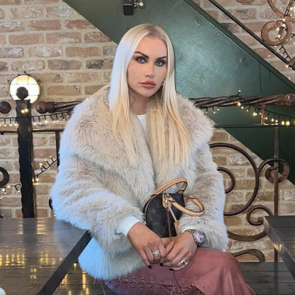 Eine Frau mit blonder, glatter Frisur trägt einen weißen Pelzmantel und hält eine schwarze Handtasche. Hintergrund: Backsteinwand und Treppengeländer.