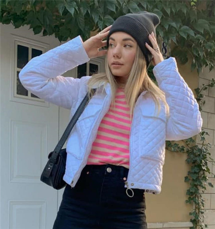 Junge Frau in gestreiftem rosa T-Shirt, weißer Steppjacke und schwarzer Mütze vor Haustür. Modetrend, Wintermode, Streetstyle.