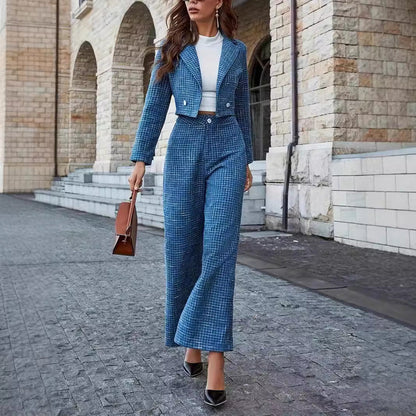 Frau in blauem Hosenanzug und weißem Oberteil, trägt braune Handtasche, geht auf gepflastertem Weg vor historischer Steinarchitektur.