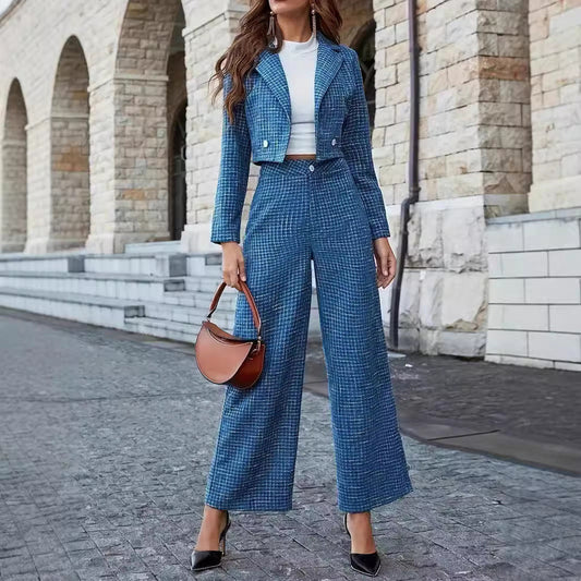 Frau in blauem Hosenanzug und weißem Top, hält braune Handtasche. Steht vor Steinmauer, trägt schwarze High Heels. Elegante Mode, urbane Kulisse.