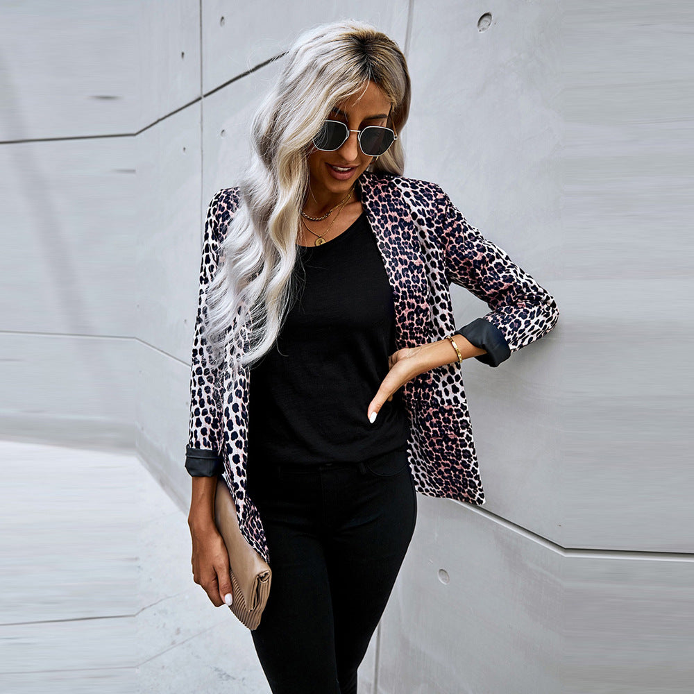 Frau mit blonder, welliger Frisur trägt Sonnenbrille, Leopardenmuster-Blazer, schwarzes Shirt und Hose, hält beige Clutch vor moderner Betonwand.