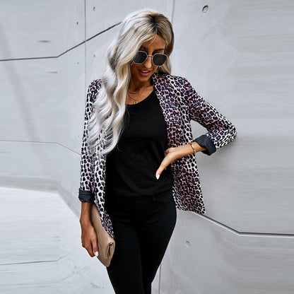 Frau mit blonder, welliger Frisur trägt Sonnenbrille, Leopardenmuster-Blazer, schwarzes Shirt und Hose, hält beige Clutch vor moderner Betonwand.