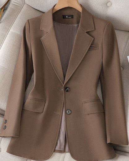 Brauner Damenblazer auf einem beigen Sessel, klassischer Schnitt mit zwei Knöpfen, Reverskragen und Pattentaschen. Elegante Businessmode.