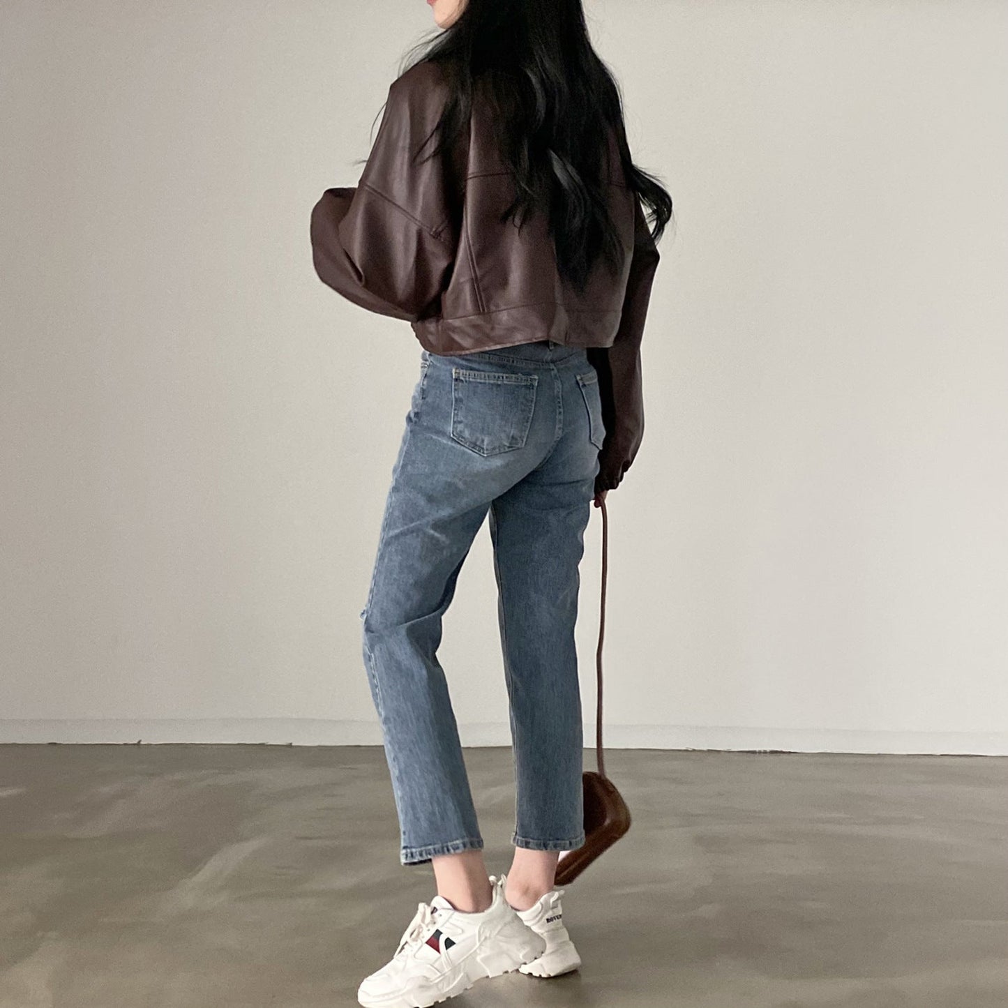 Frau in brauner Lederjacke und blauer Jeans, trägt weiße Sneakers, steht auf Betonboden. Modetrend, lässiger Stil, Herbstmode.