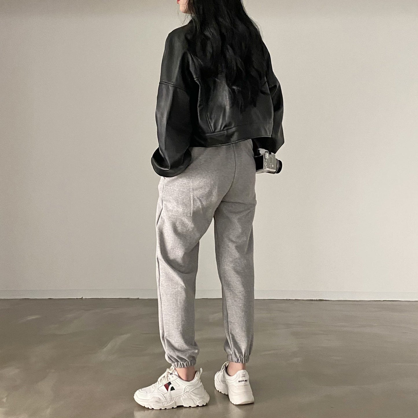 Frau in schwarzer Lederjacke und grauer Jogginghose, weiße Sneaker, minimalistische Mode, lässiger Stil, Streetwear, Rückansicht.