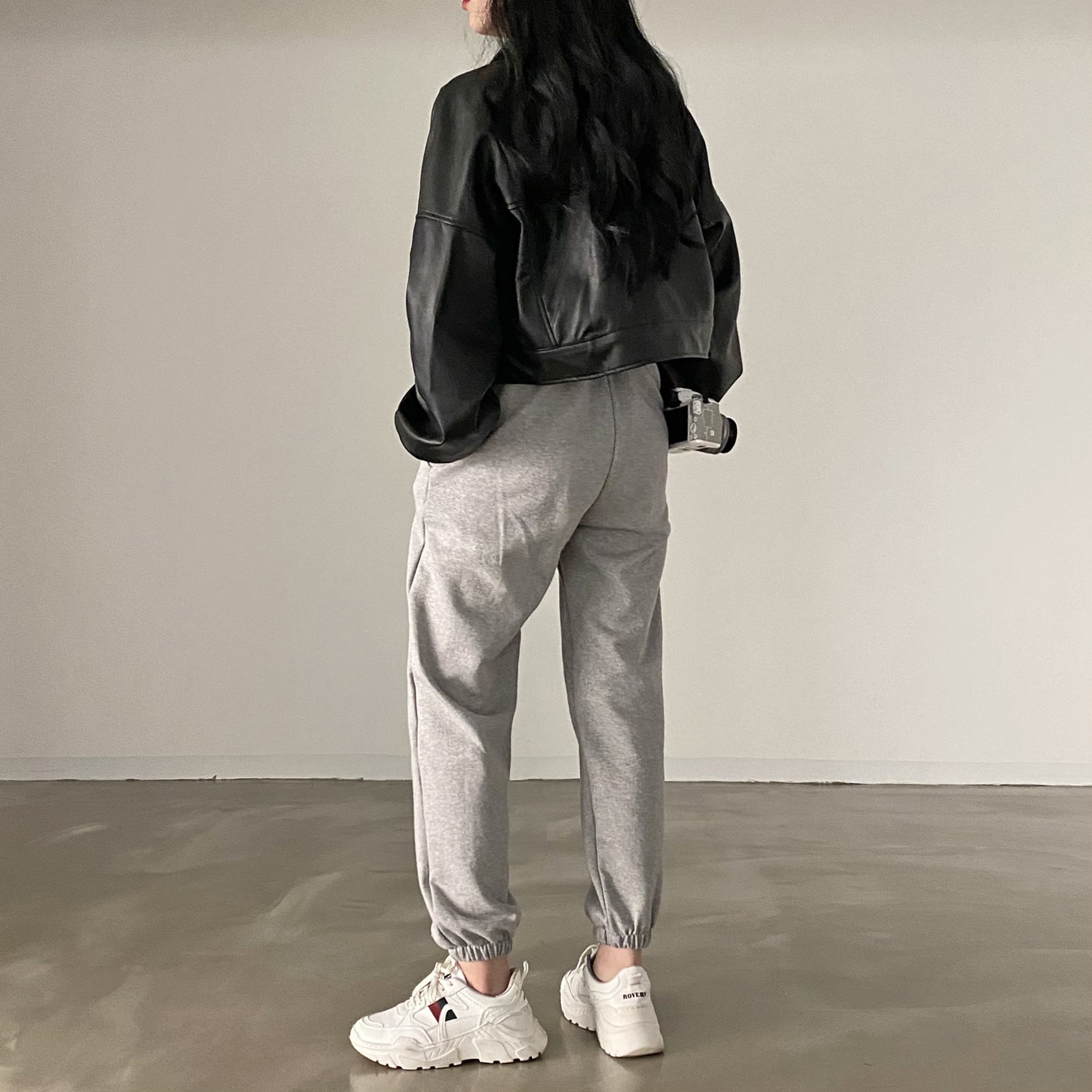 Frau in schwarzer Lederjacke und grauer Jogginghose, weiße Sneaker, minimalistische Mode, lässiger Stil, Streetwear, Rückansicht.
