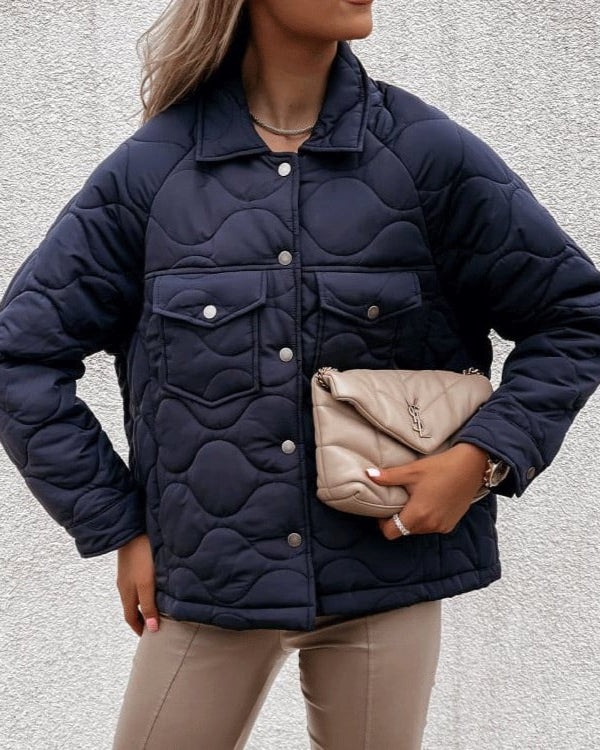 Frau in dunkelblauer Steppjacke mit beiger Designer-Clutch vor weißer Wand. Modetrend, Herbstmode, Damenjacke, stilvolles Outfit.