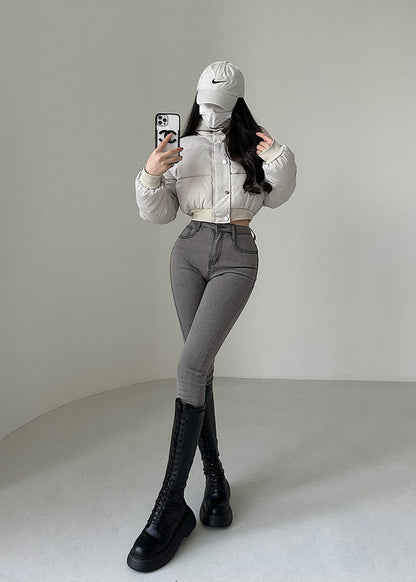 Frau in modischer Winterkleidung mit weißer Daunenjacke, grauer Hose, schwarzen Stiefeln und Baseballkappe, macht Selfie vor weißem Hintergrund.