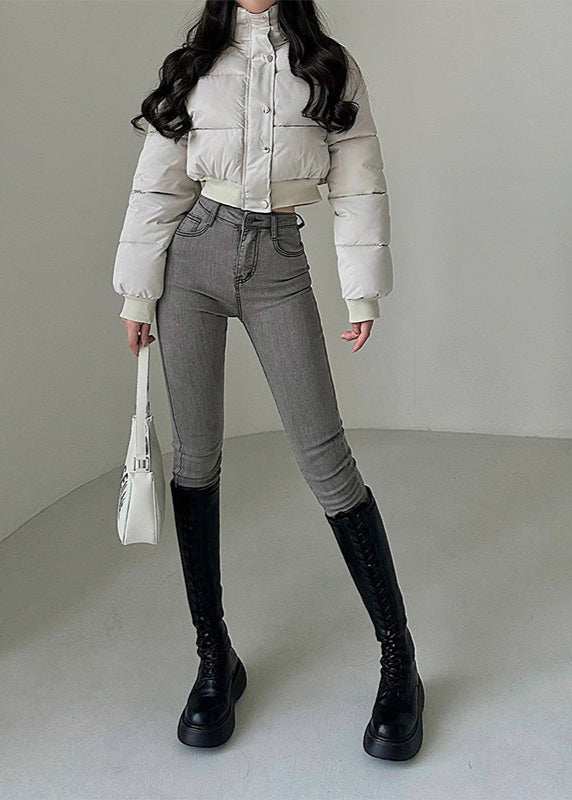 Frau in weißer Daunenjacke, grauer Jeans und schwarzen Stiefeln, hält weiße Handtasche. Modetrend, Winteroutfit, stilvolle Damenbekleidung.