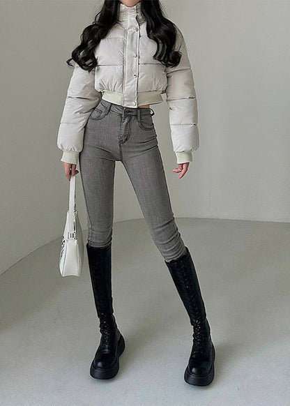 Frau in weißer Daunenjacke, grauer Jeans und schwarzen Stiefeln, hält weiße Handtasche. Modetrend, Winteroutfit, stilvolle Damenbekleidung.