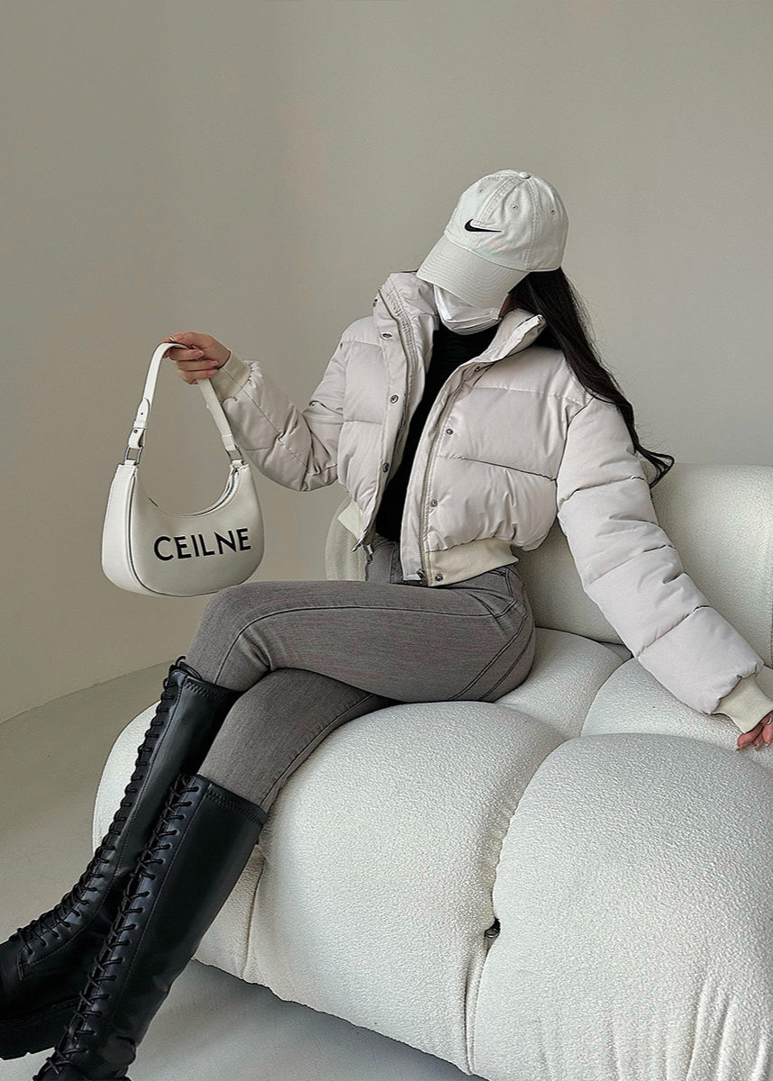 Frau in weißer Daunenjacke und Baseballkappe sitzt auf Sofa, hält Designer-Handtasche. Modetrend, Wintermode, stilvolle Damenbekleidung.