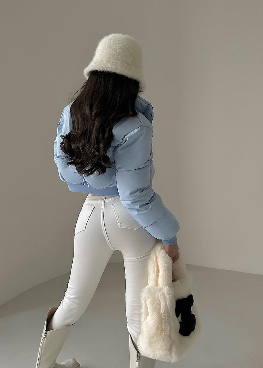 Frau in blauer Daunenjacke, weißer Hose und Stiefeln, hält flauschige Tasche. Wintermode, stilvoller Look, modische Kleidung.