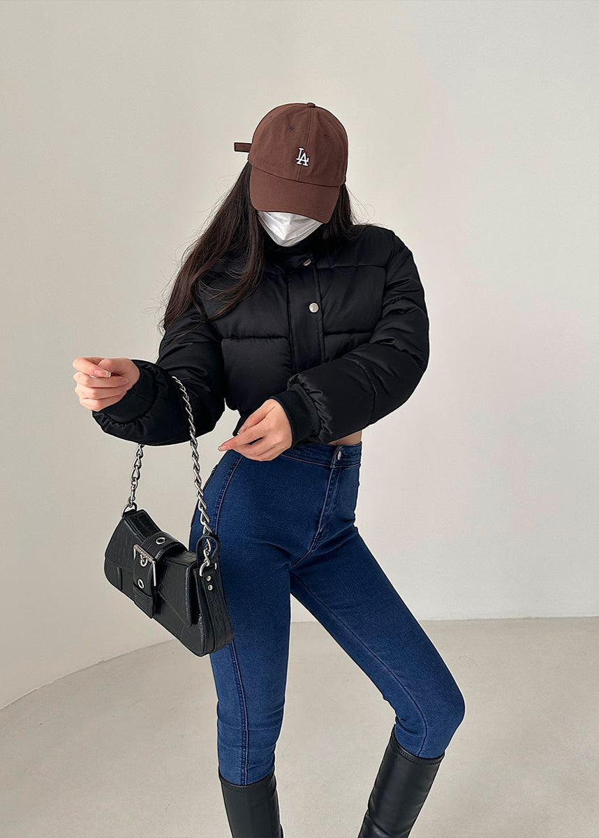 Frau in schwarzer Daunenjacke, blauer Jeans und brauner Kappe, hält schwarze Handtasche. Modetrend, Winteroutfit, Streetstyle.