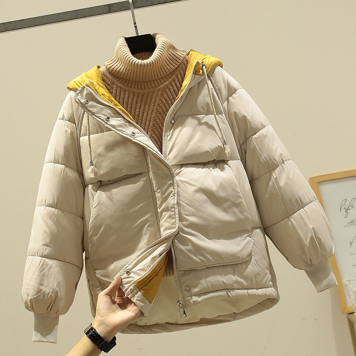 Beige Daunenjacke mit gelbem Innenfutter und Kapuze, über einem beigen Rollkragenpullover. Wintermode, warme Oberbekleidung, stilvoll und gemütlich.