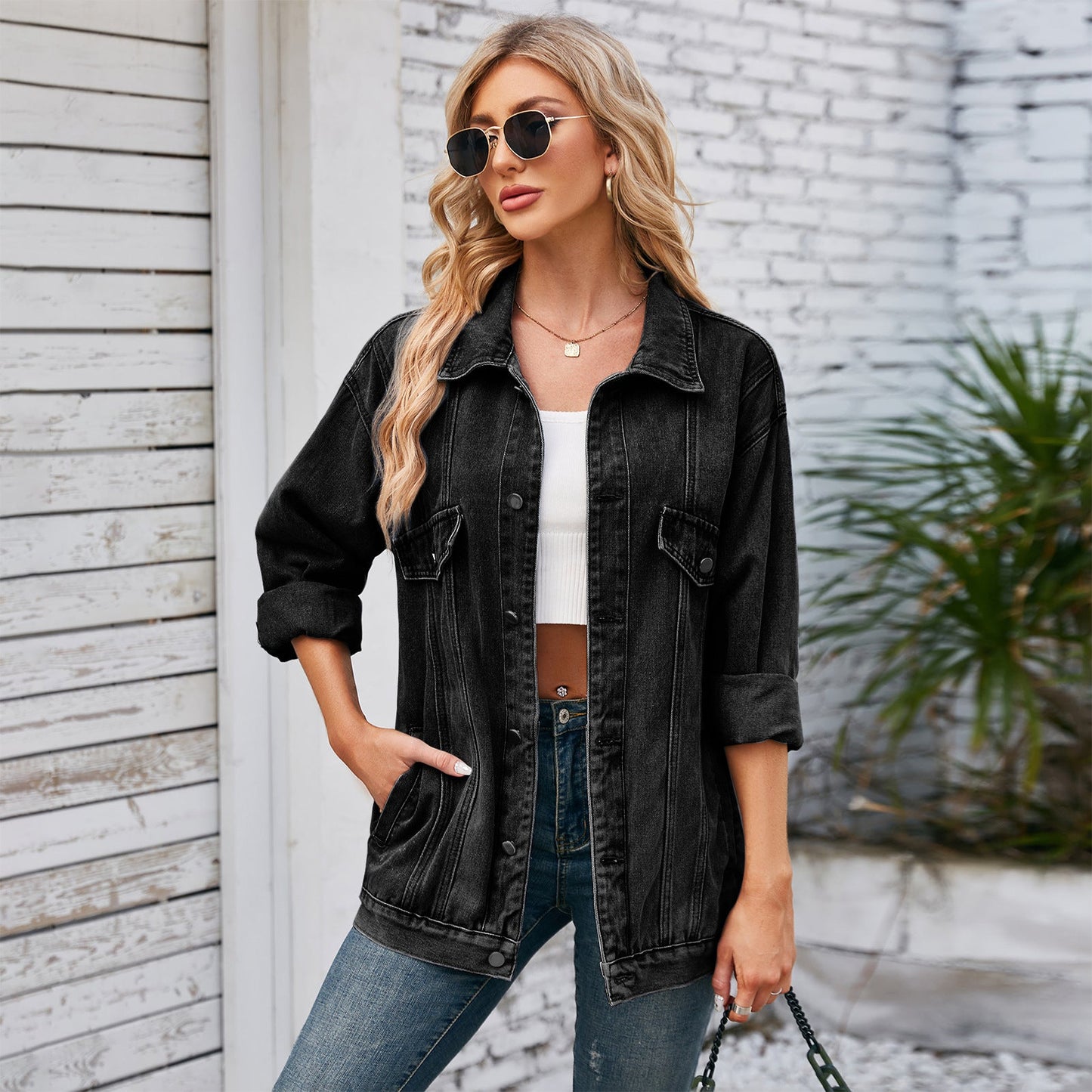 Blonde Frau in schwarzer Jeansjacke, weißem Top und blauer Jeans, trägt Sonnenbrille, steht vor weißer Backsteinwand. Trendige Damenmode, lässiger Look.