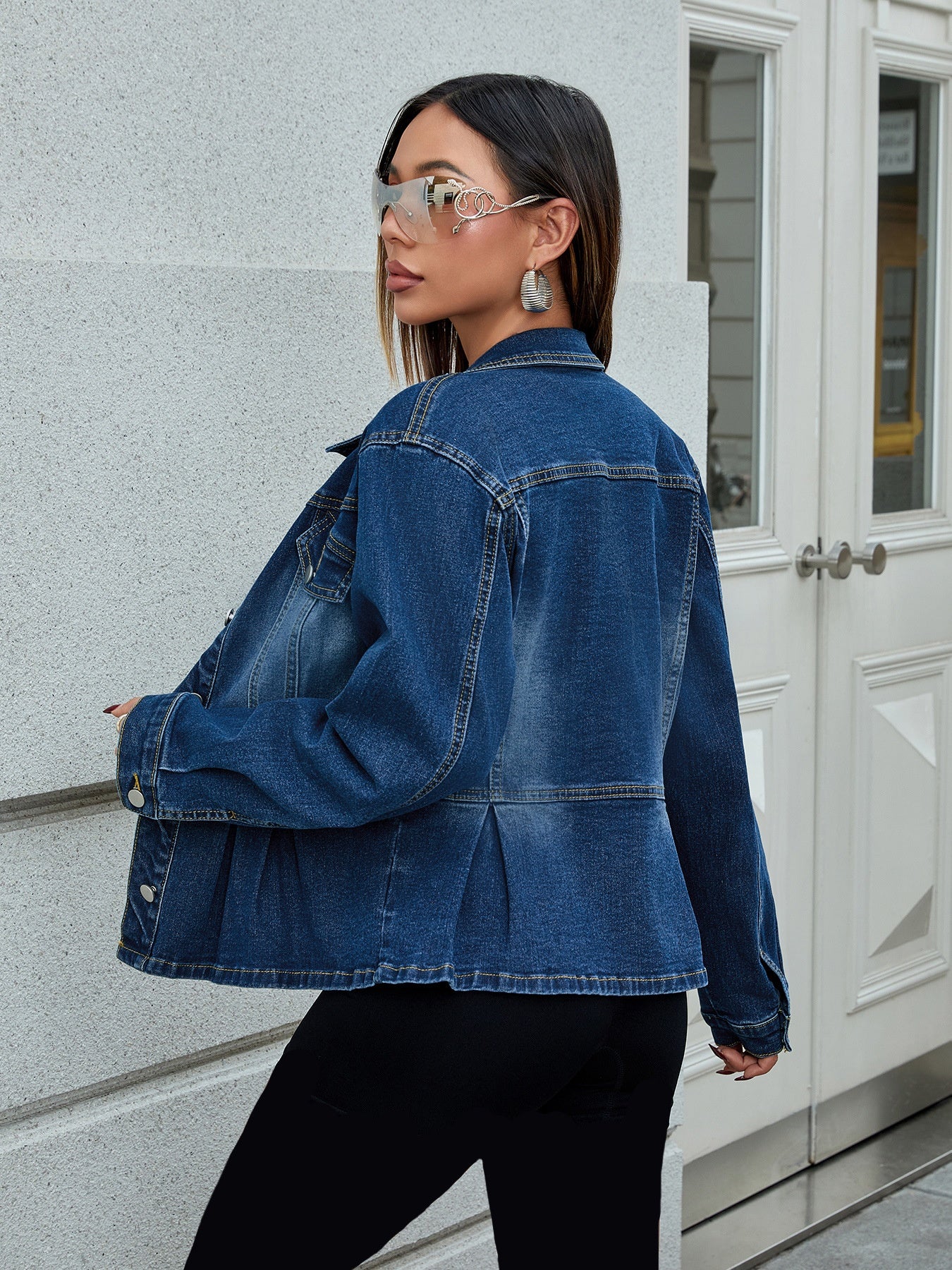 Frau in blauer Jeansjacke und Sonnenbrille, steht vor weißer Tür. Modetrend, Streetstyle, lässiger Look, Damenmode, urbaner Stil.
