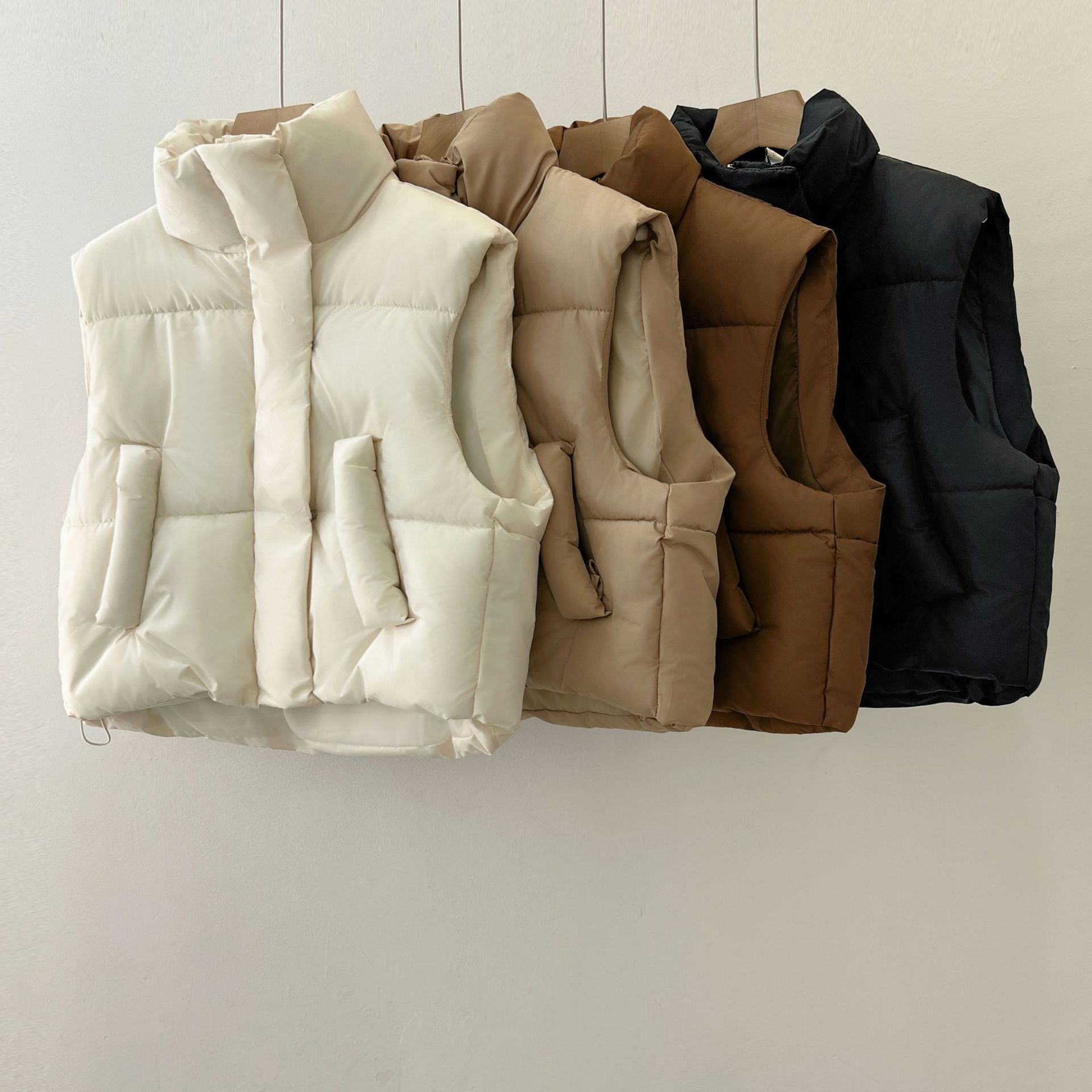 Vier gesteppte Daunenwesten in Weiß, Beige, Braun und Schwarz hängen an Haken. Modische Winterbekleidung, warm, ärmellos, vielseitig.