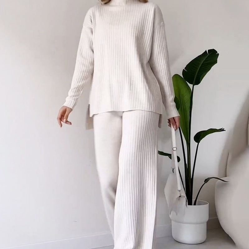 Frau in cremefarbenem Strickset mit Pullover und Hose, hält weiße Tasche. Minimalistisches Outfit, moderne Mode, Zimmerpflanze im Hintergrund.