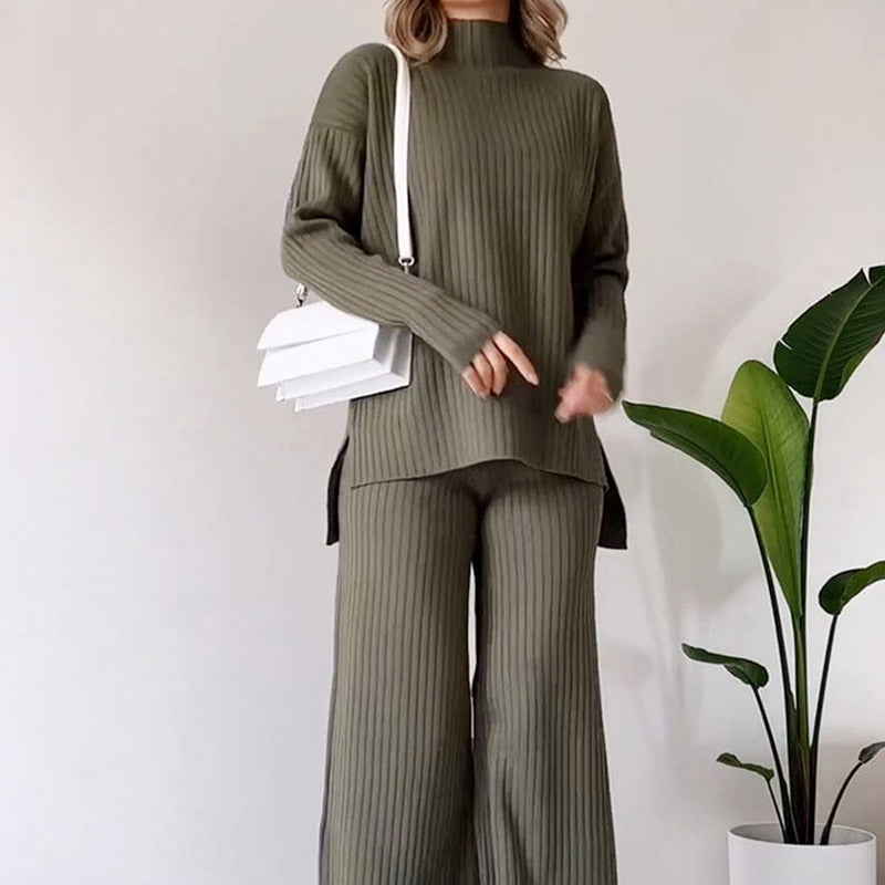 Frau in olivgrünem, geripptem Strick-Zweiteiler mit weißer Umhängetasche, steht vor einer Zimmerpflanze. Modetrend, Herbstmode, Damenbekleidung.