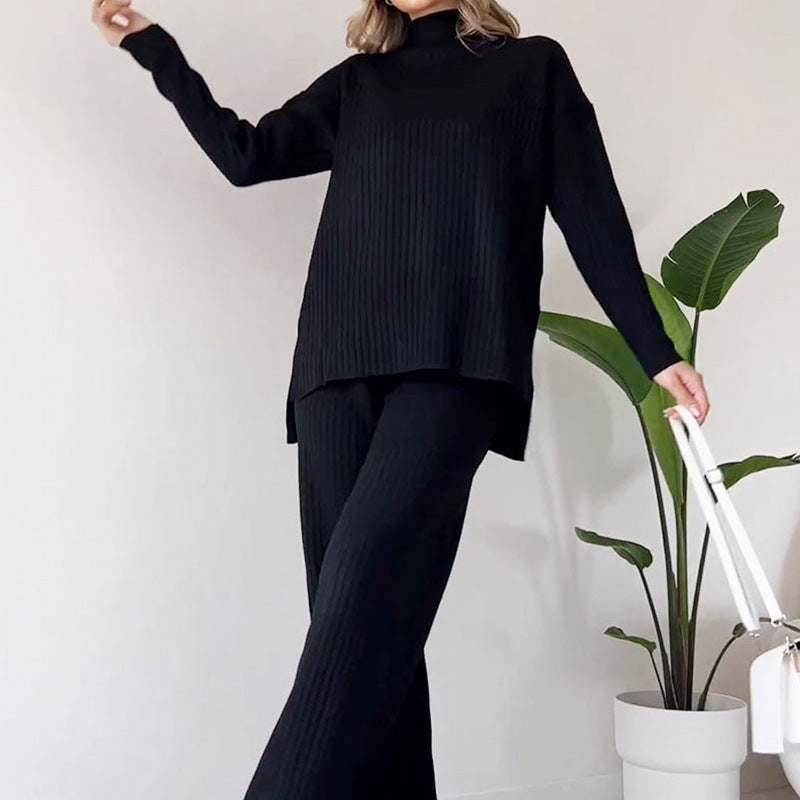Frau in schwarzem Strickpullover und Hose posiert neben einer Pflanze. Modetrend, Herbstmode, eleganter Look, minimalistisch, gemütlich.