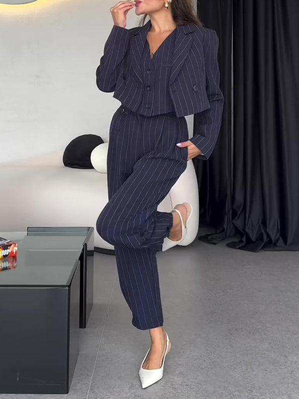 Frau in dunkelblauem Nadelstreifenanzug, posiert stilvoll im modernen Wohnzimmer. Eleganter Business-Look, weiße Schuhe, schwarze Vorhänge im Hintergrund.