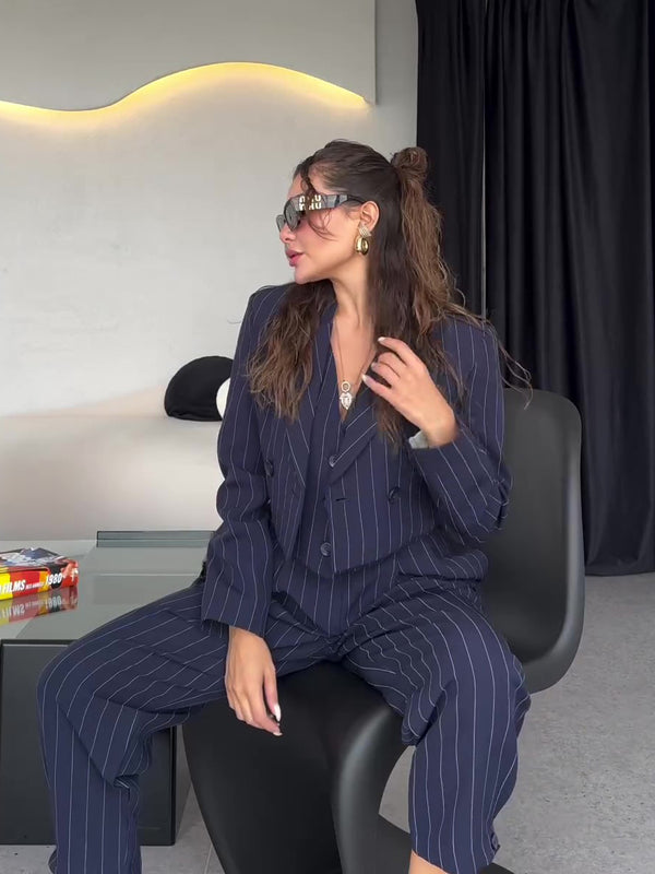 Frau in gestreiftem Anzug sitzt auf modernem Stuhl, trägt Sonnenbrille und Schmuck. Stilvolle Businessmode, elegantes Büro-Outfit, trendige Damenbekleidung.