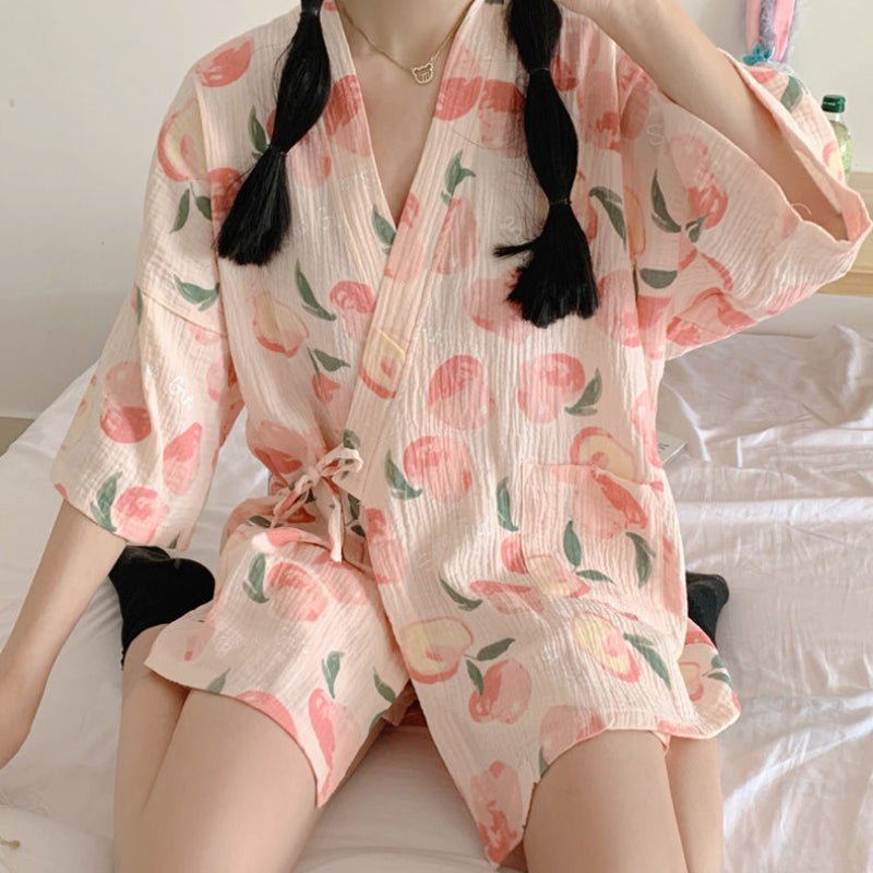Person in einem pfirsichgemusterten Kimono sitzt auf einem Bett. Der Kimono hat kurze Ärmel und einen Bindegürtel. Ideal für Sommermode und Freizeitkleidung.