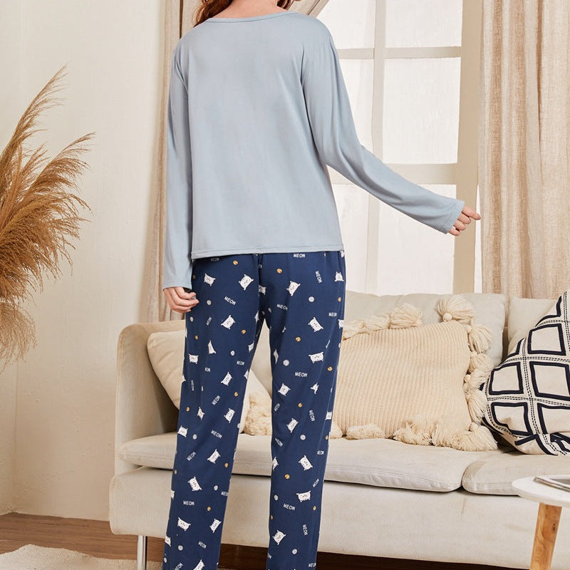 Frau in blauer Pyjama-Hose mit Muster und grauem Langarmshirt steht im Wohnzimmer. Gemütliche Einrichtung mit Sofa und Kissen im Hintergrund.