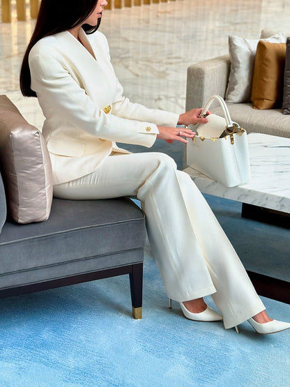 Elegante Frau im weißen Anzug sitzt auf einem Sofa, hält eine weiße Designer-Handtasche. Luxuriöse Innenausstattung, stilvolle Mode, Business-Look.