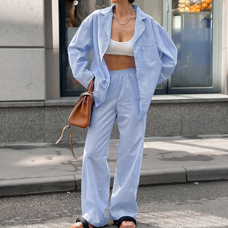 Frau in hellblauem Freizeitanzug und weißem Top, trägt braune Tasche, steht auf Bürgersteig vor Schaufenster. Modetrend, lässiger Stil.