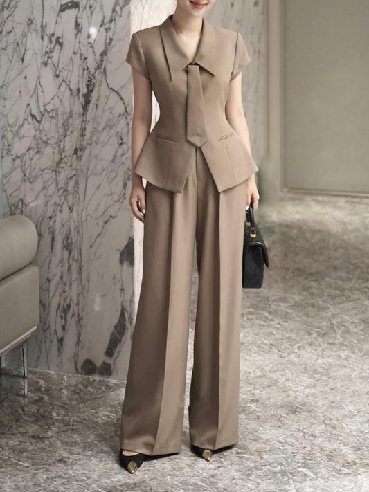 Eleganter Damenanzug in Beige, bestehend aus tailliertem Blazer und weiten Hosen, kombiniert mit schwarzen Schuhen und Handtasche, vor Marmorkulisse.