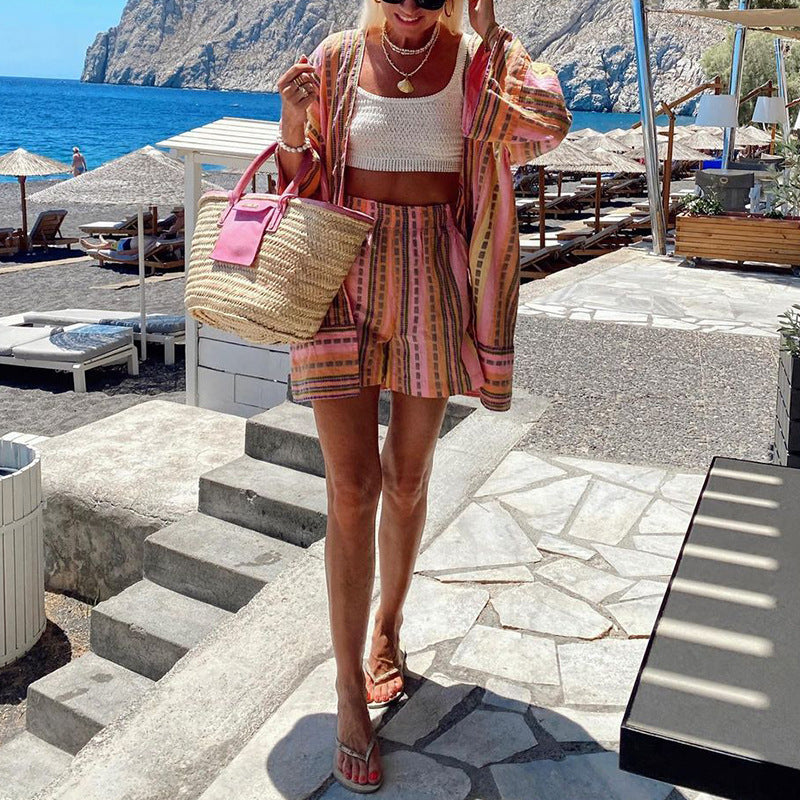 Frau in gestreiftem Sommeroutfit mit Strohtasche am Strand, Meer und Liegestühle im Hintergrund, sonniger Tag, Urlaubsstimmung.