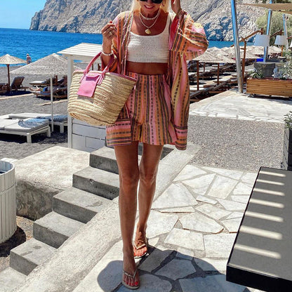 Frau in gestreiftem Sommeroutfit mit Strohtasche am Strand, Meer und Liegestühle im Hintergrund, sonniger Tag, Urlaubsstimmung.