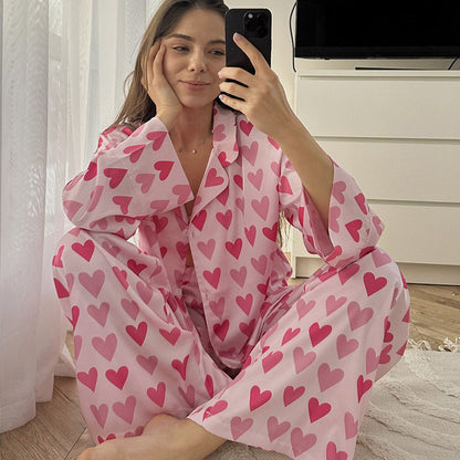 Frau in rosa Pyjama mit Herzmuster, sitzt lächelnd auf dem Boden und macht ein Selfie. Schlafzimmer im Hintergrund. Bequeme Nachtwäsche, Selfie.