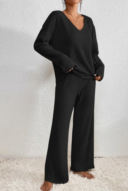 Frau in schwarzem Strickpullover und Hose, gemütliches Lounge-Set, modisches Herbst-Winter-Outfit, lässiger Stil, auf weißem Teppich stehend.
