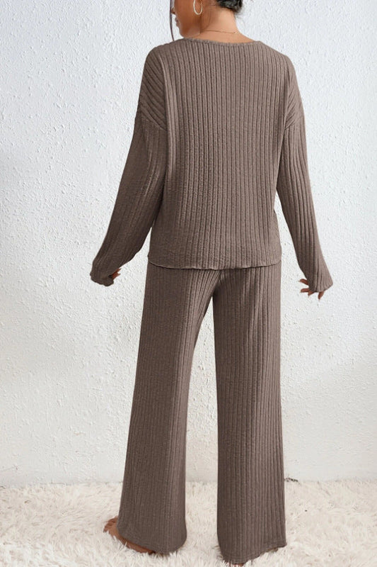 Frau in braunem, geripptem Strick-Zweiteiler, bestehend aus Pullover und Hose, vor weißer Wand. Bequemes, modisches Loungewear-Set.