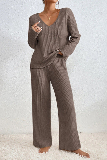 Frau in braunem Strick-Zweiteiler, bestehend aus V-Ausschnitt-Pullover und weiten Hosen, steht barfuß auf weißem Teppich vor weißer Wand.