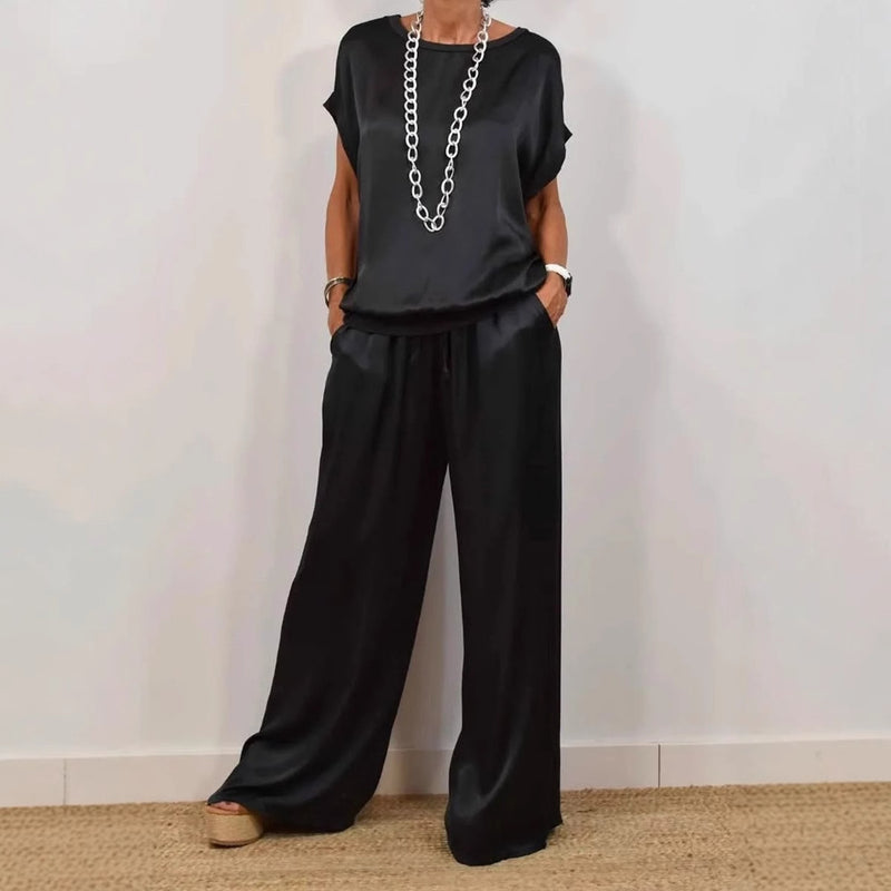 Frau in schwarzem Satin-Zweiteiler mit weitem Schnitt, bestehend aus T-Shirt und Hose, trägt lange silberne Kette, modisch, elegant, lässig.