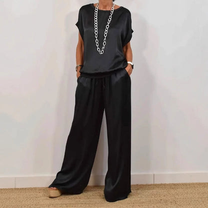 Frau in schwarzem Satin-Zweiteiler mit weitem Schnitt, bestehend aus T-Shirt und Hose, trägt lange silberne Kette, modisch, elegant, lässig.