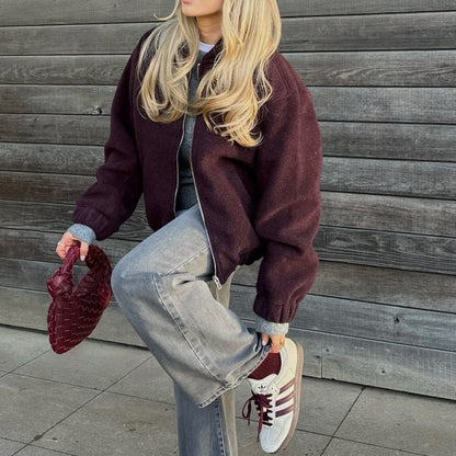 Blonde Frau in lila Jacke, grauer Jeans und Adidas-Sneakern, hält rote Tasche. Streetstyle-Mode, lässiger Look, Holzwand im Hintergrund.