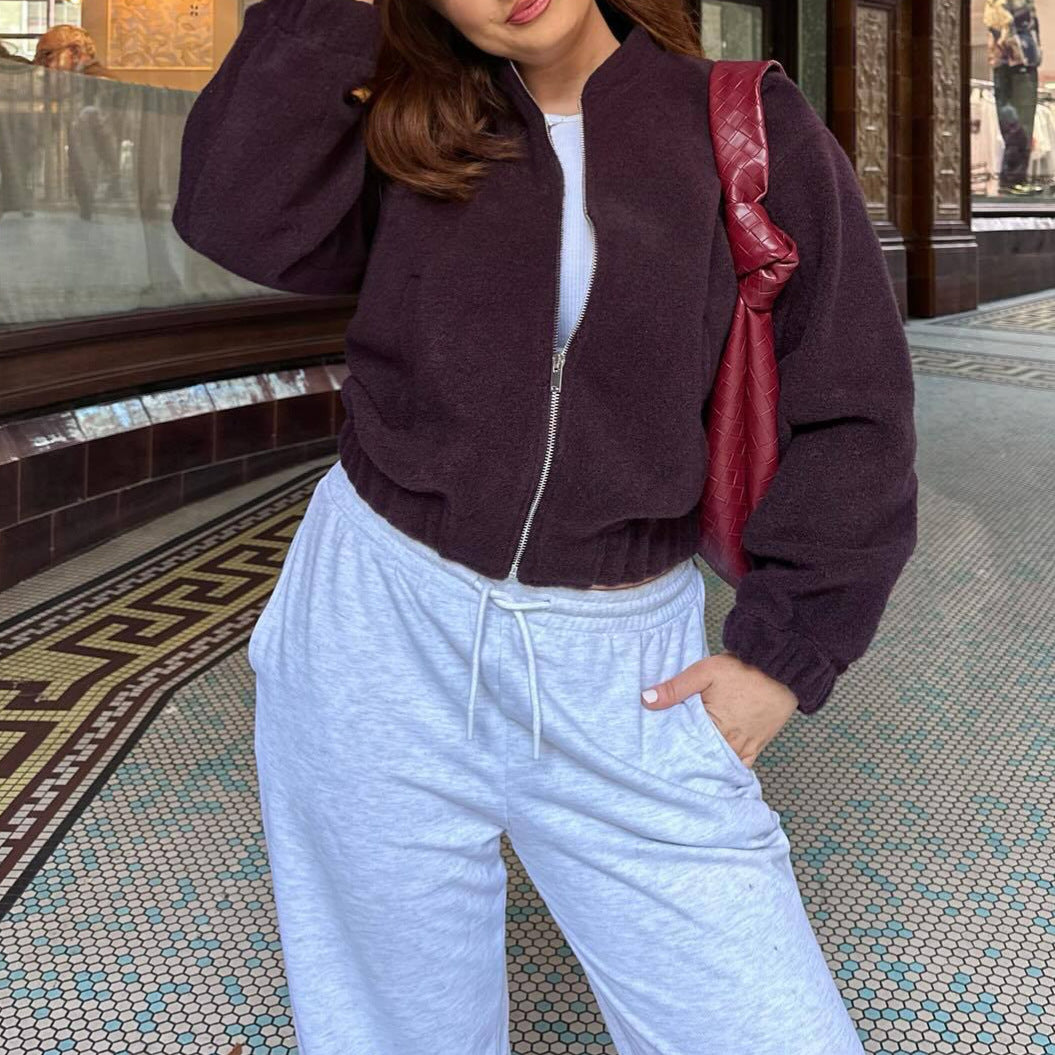 Frau in lila Bomberjacke und grauer Jogginghose, roter Tasche, steht auf gemustertem Boden. Modetrend, lässiger Stil, urbane Kleidung.